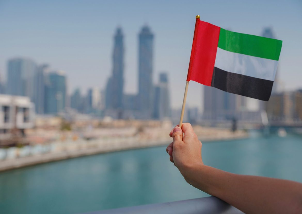 female hand holding tiny flag of UAE against Dubai skyline.