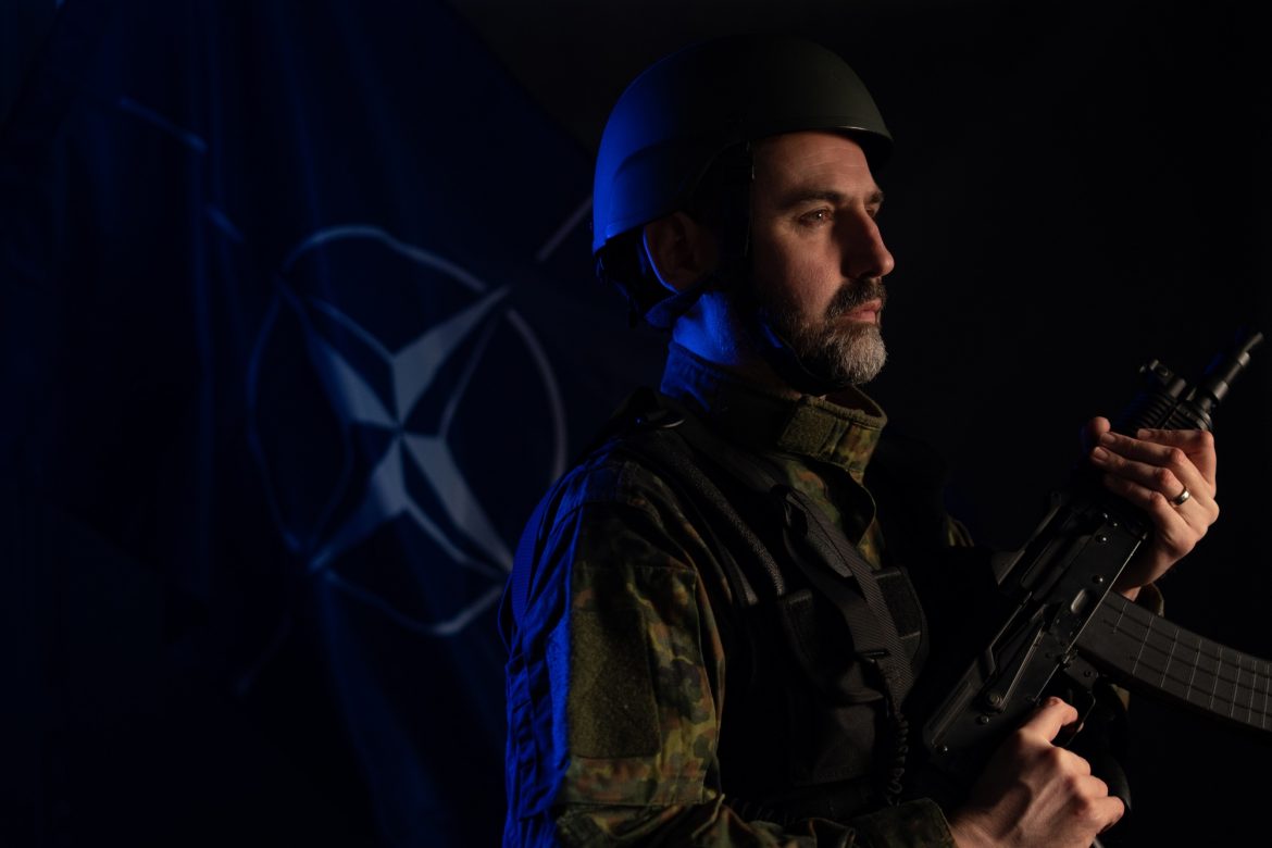 Soldier in military uniform and helmet holding weapon with NATO flag in background
